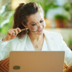 smiling trendy 40 years old woman in white blouse and jacket with laptop using magnifying glass in the modern living room in sunny day. (smiling trendy 40 years old woman in white blouse and jacket with laptop using magnifying glass in the modern livi